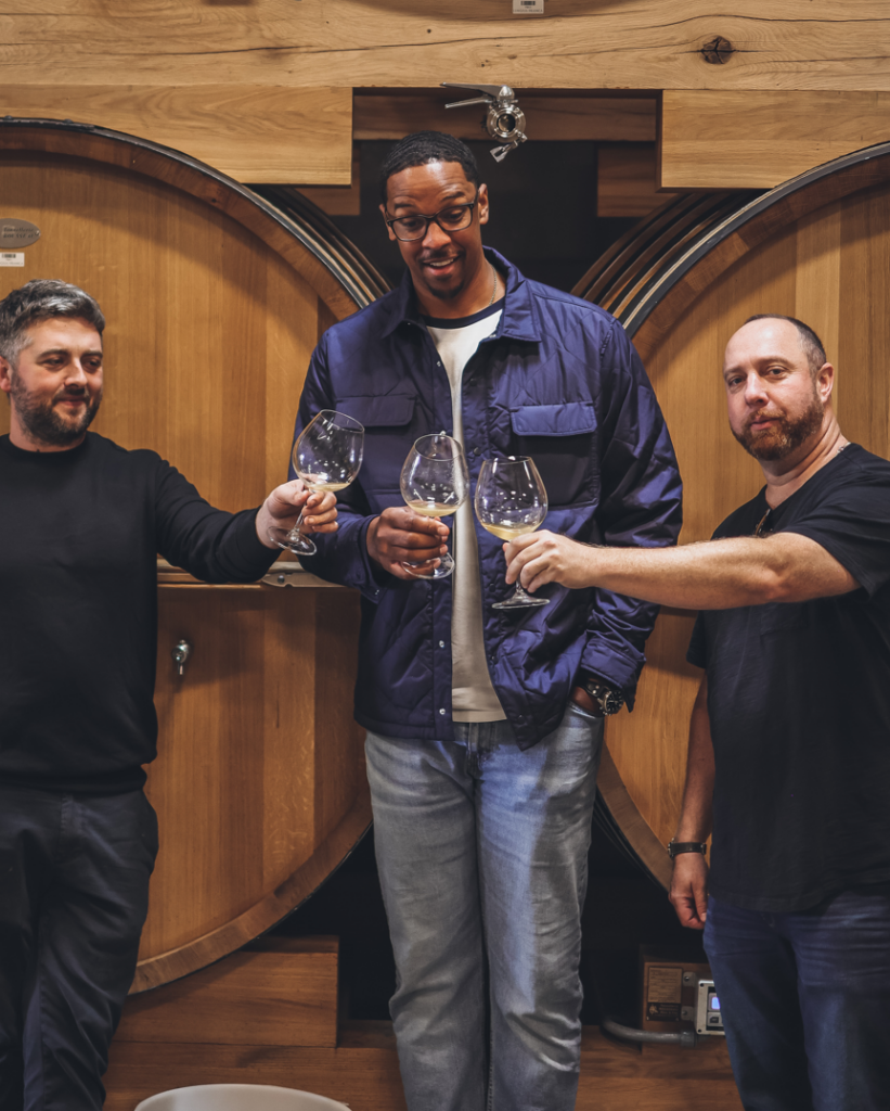 Thomas Savre, Director of Winemaking at Lingua Franca, Channing Frye, owner at Chosen Family Wines and Jacob Gray, Partner at Chosen Family Wines, toasting in front of wine barrels.